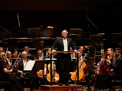 A conductor smiling on stage with the orchestra behind him.