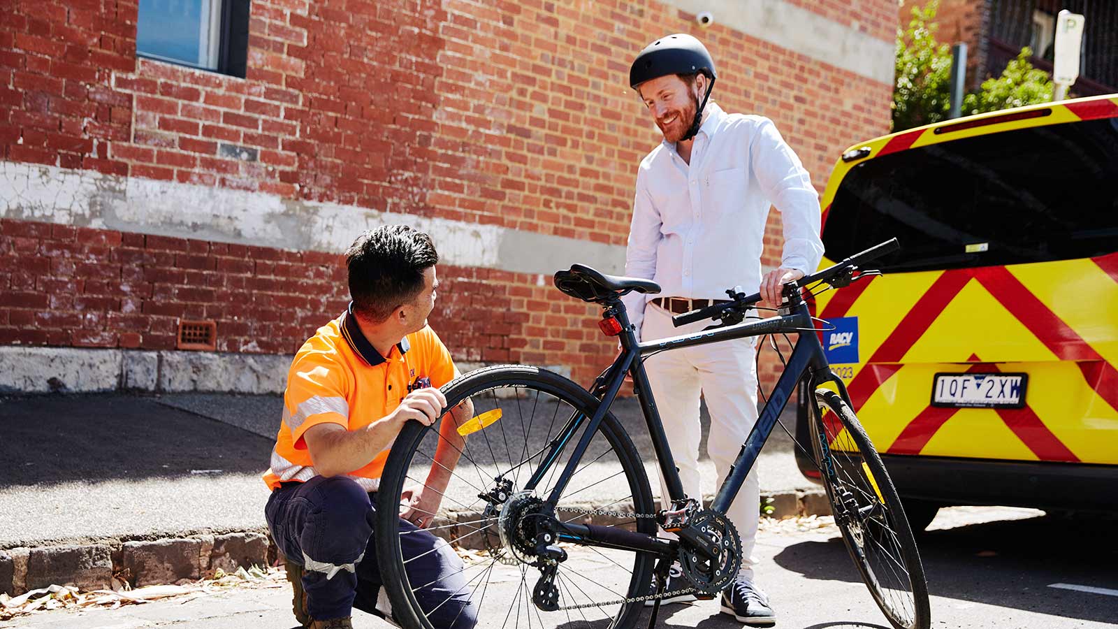RACV technician assessing a bike and speaking with a smiling customer, with the RACV yellow patrol van behind them.