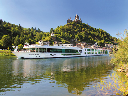 A river cruise boat going by a european riverside town with a castle on a hill in the background.