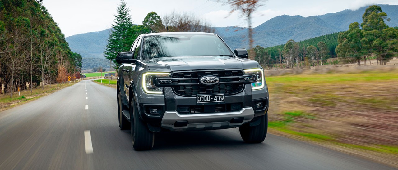 2024-Ford-Ranger-Tremor-banner-1400x600 Metallic grey Ford Ranger Tremor dual-cab ute on rural tree-lined bitumen road with high country behind