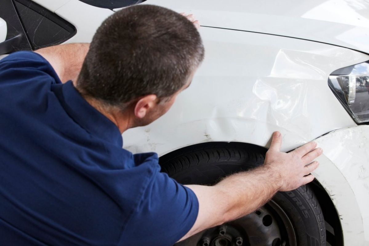 A panel beater inspects a dent on the front bumper of a white sedan-type car