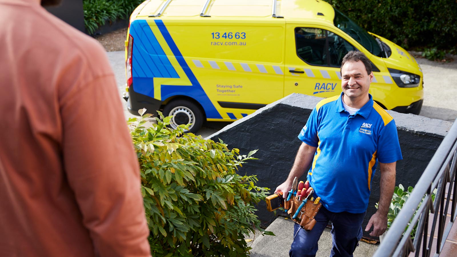 RACV Tradesman smiling and greeting homeowner on front porch with yellow and blue tradie van in background.