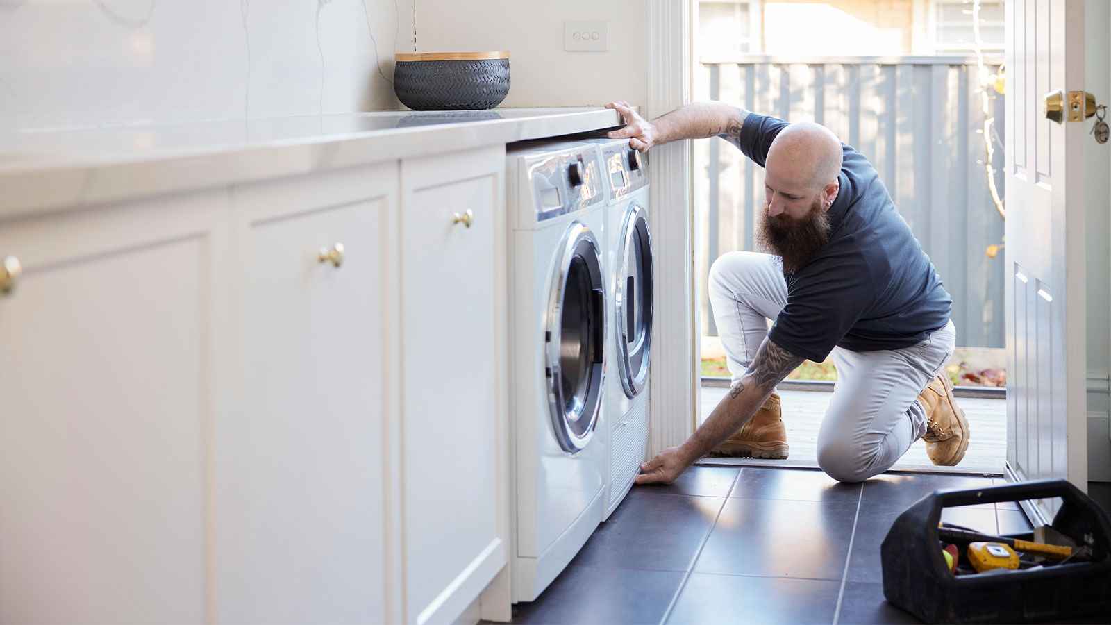 Tradesperson kneals to fix washing machine at a customer's house.