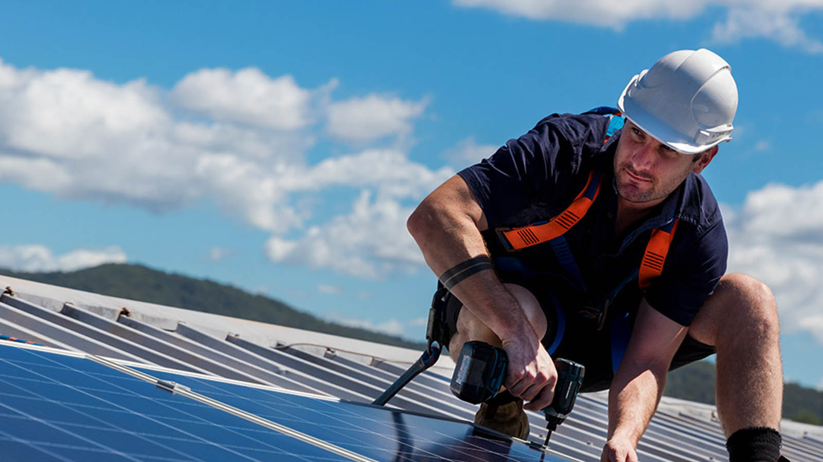 Tradesperson wearing PPE on a roof installing solar panelling with a drill.