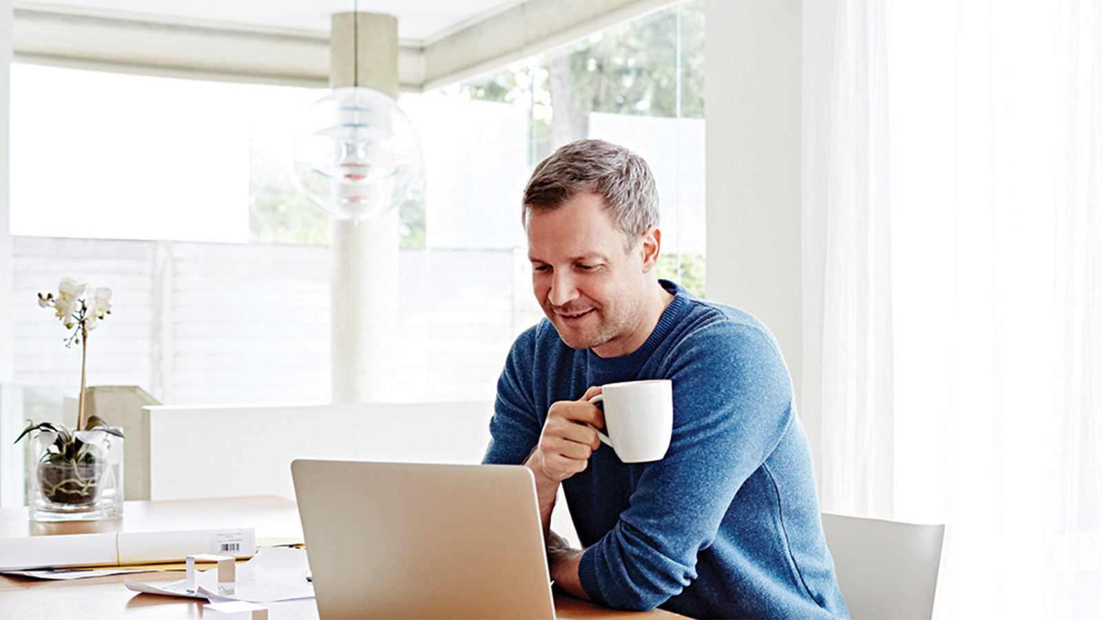 Man looking at his laptop and holding a white mug, in front of sheer curtains
