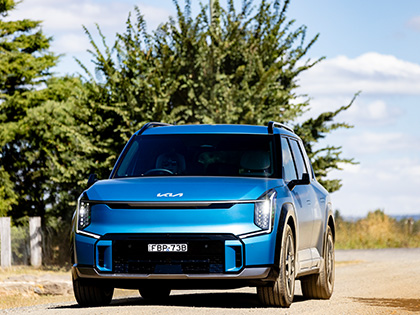 Blue Kia EV9 on a road with green trees and the blue sky behind.