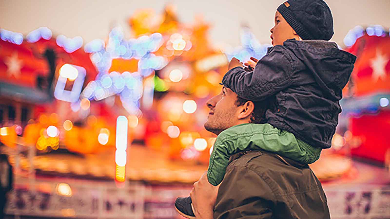 Child sitting on father's shoulders at a carnival, with red and yellow lights out of focus in the background.