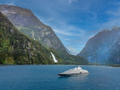 A small cruise boat floating near a lush waterfall in Milford Sound on a blue skied day.