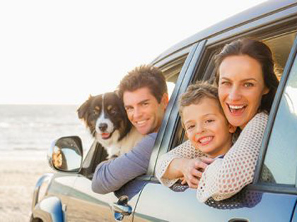 A young family and a dog smiling outside of a car window whilst at the beach.