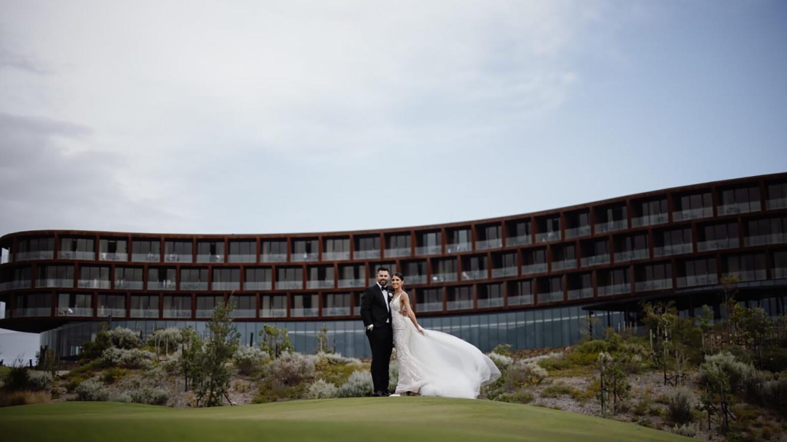 Couple posing on grass in front of RACV Cape Schanck resort, wearing wedding dress and suit.