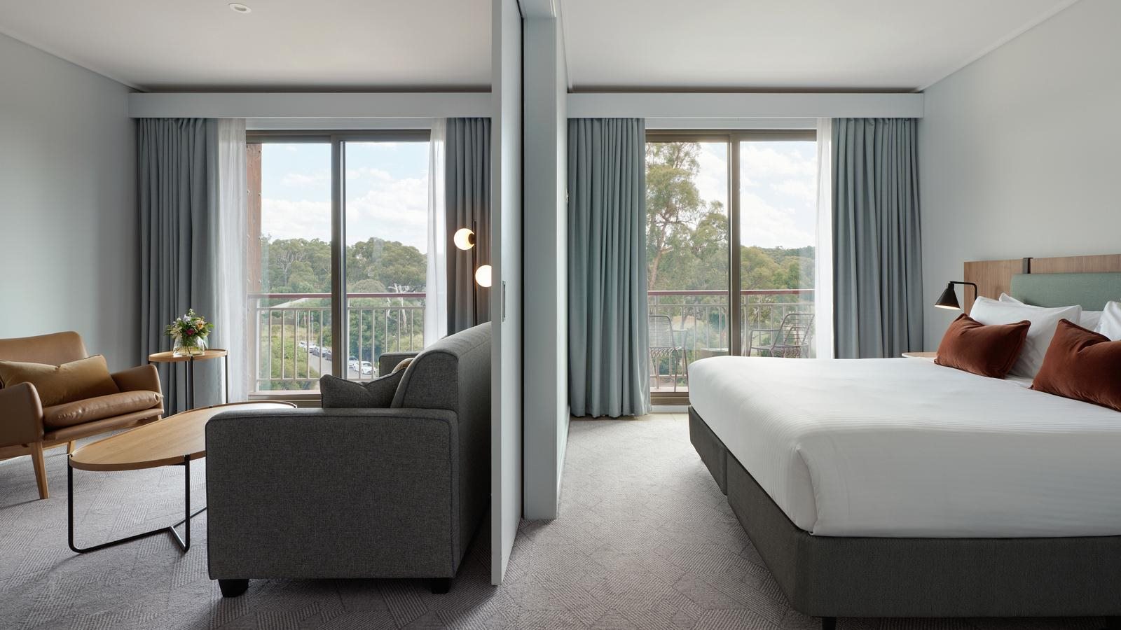 Goldfields studio room with a large bed, a sofa, and a table. The room has two large windows with curtains and a view of greenery outside.