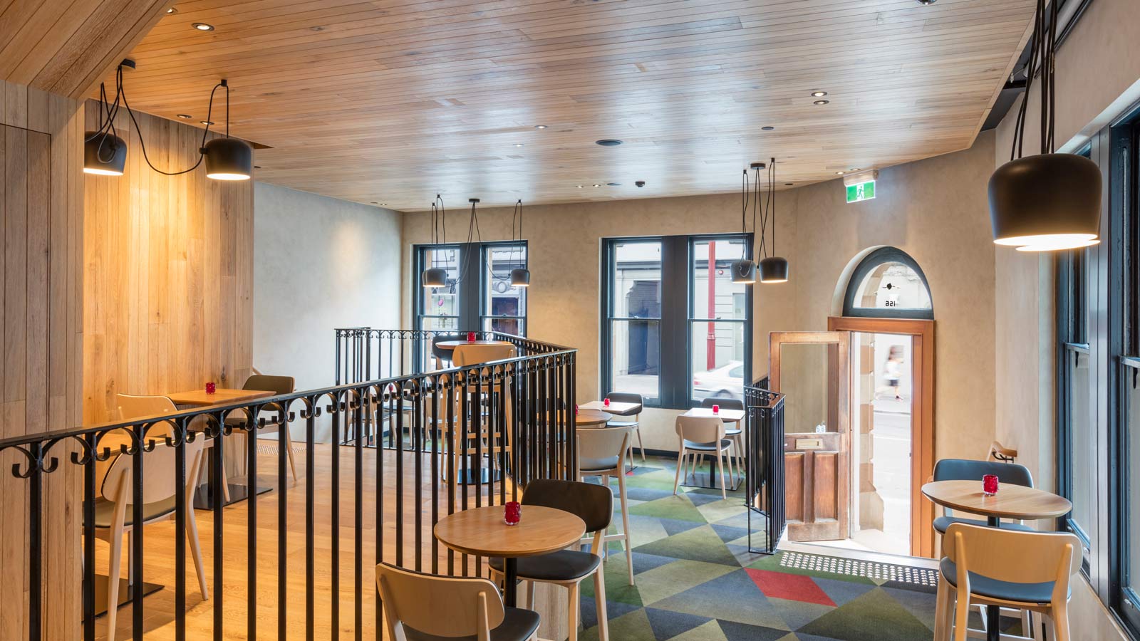 Interior view of RACV Hobart Hotel's Cascade on Collins bar, featuring wooden flooring and ceiling panels, metal railing separating different sections, and a mixture of warm lighting from styling pendant lamps and natural light coming through the large windows in the background.