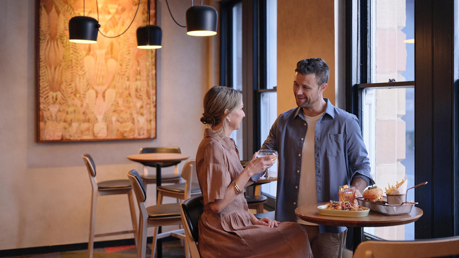 Couple smiling together by the window at RACV Hobart Hotel's Cascade on Collins bar, with pub style food on the table in front of them.