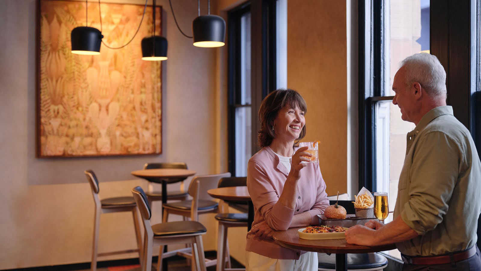 Couple smiling together by the window at RACV Hobart Hotel's Cascade on Collins bar, with pub style food on the table in front of them.