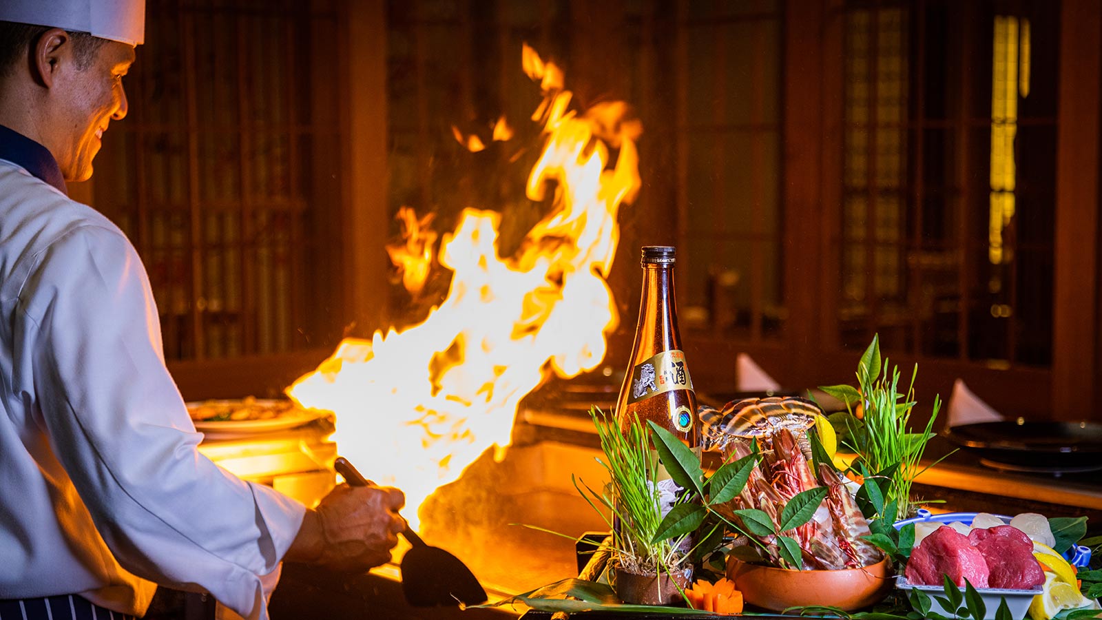 RACV Royal Pines Resort chef smiling while flambeing over a hot plate at the Arakawa restaurant, standing near fresh produce.