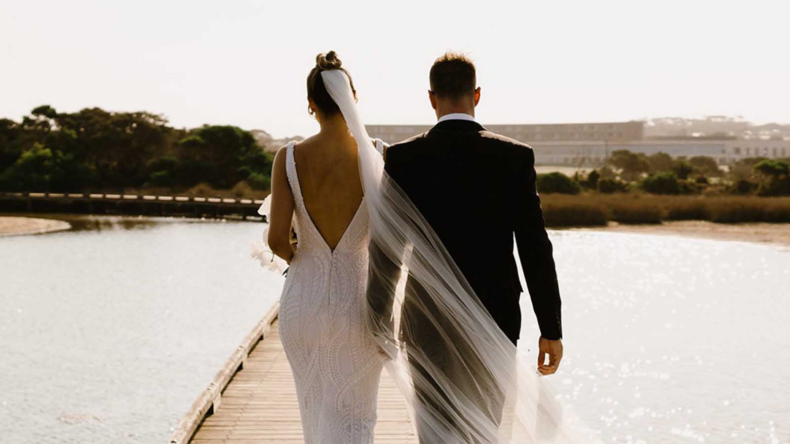 Couple walking on a pier with their backs to the camera, wearing a wedding dress and suit.