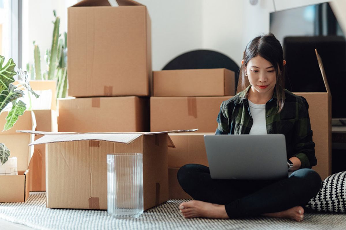 A woman working on a laptop on the ground while a stack of moving boxes sit behind her