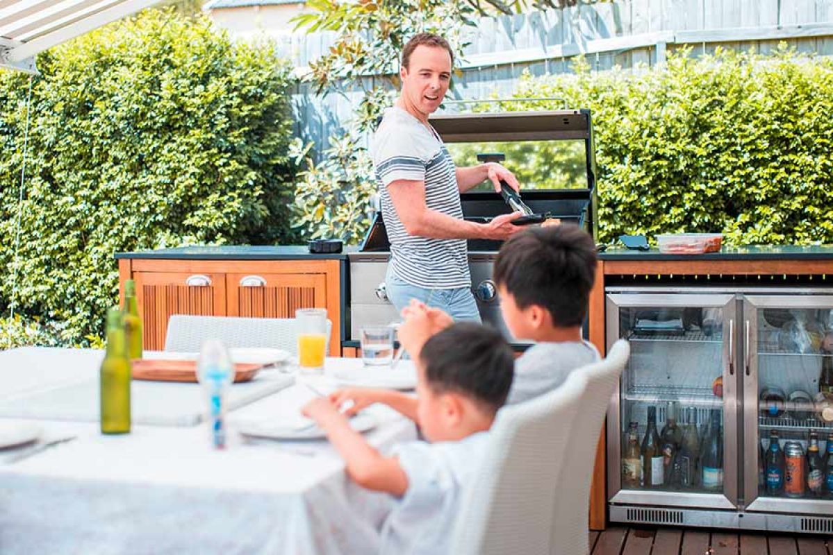 Man cooking on outdoor barbecue while kids sit at table eating lunch 