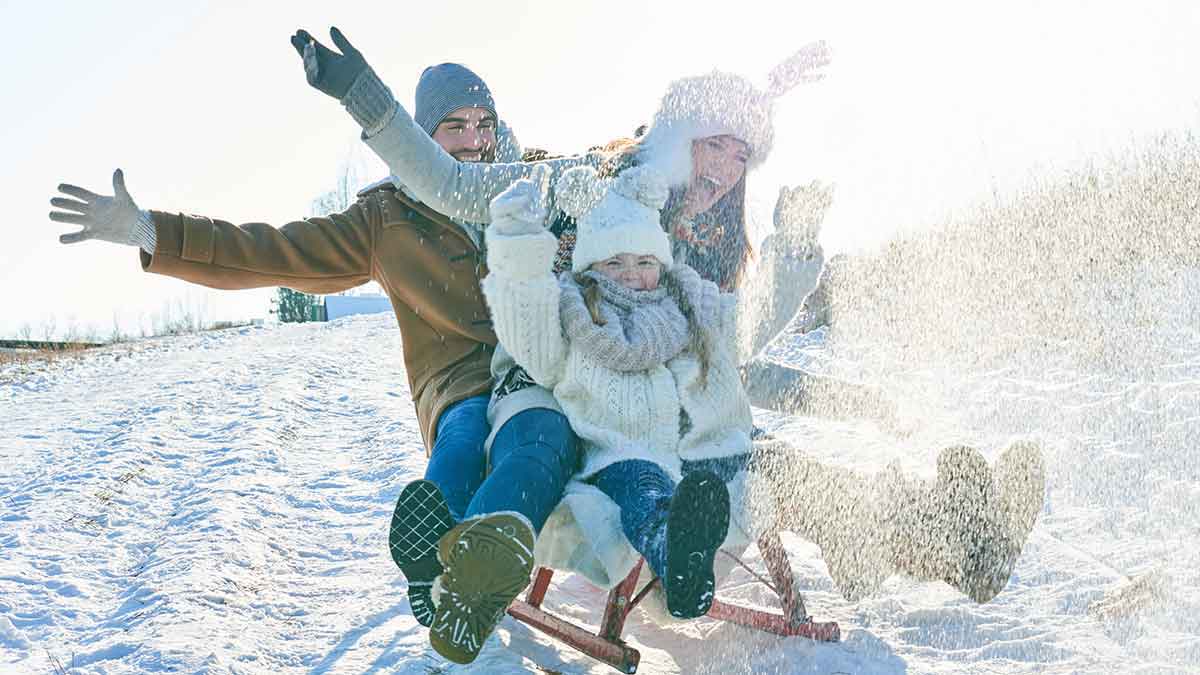 Family with their hands up in the air, tobogganing down a hill with snow spraying up