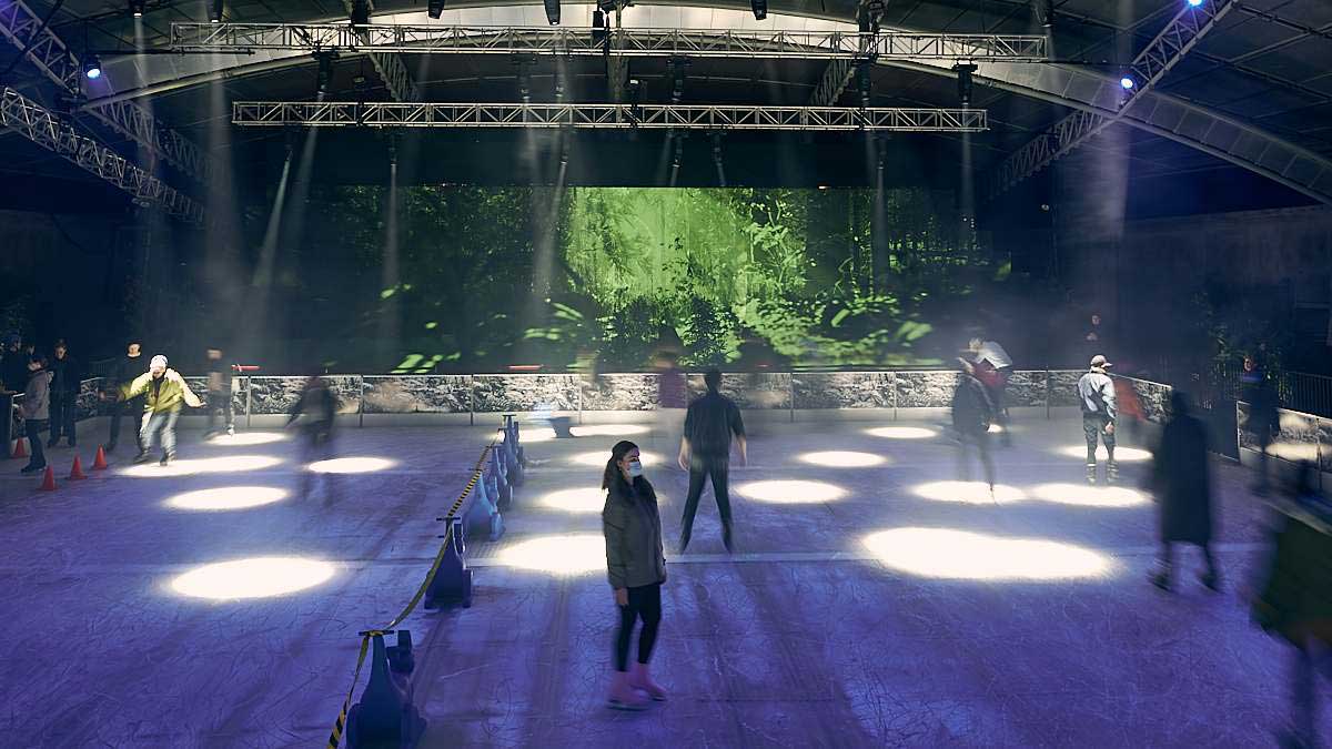 A crowd ice skating under spotlights with a projection screen showing a forest of ferns