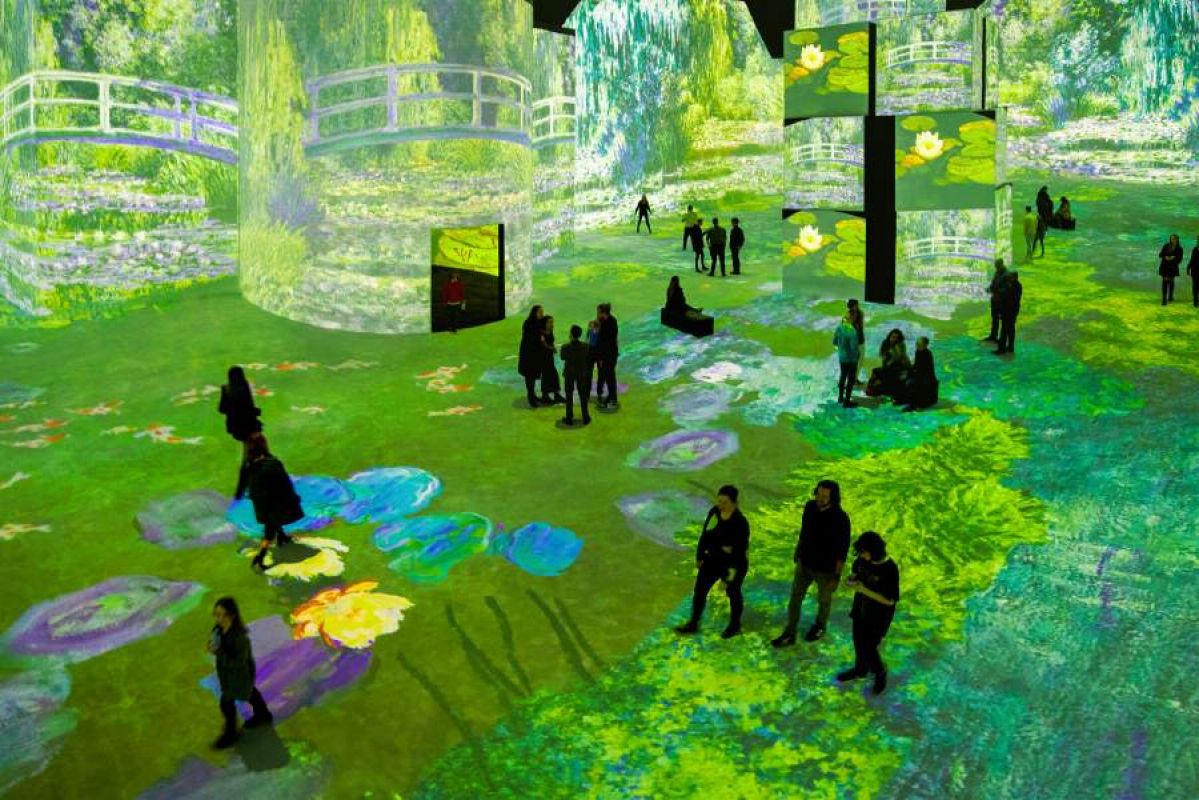 People walking through a room in which Monet's 'Bridge over a Pond of Water Lilies' is projected on every surface at the Lume