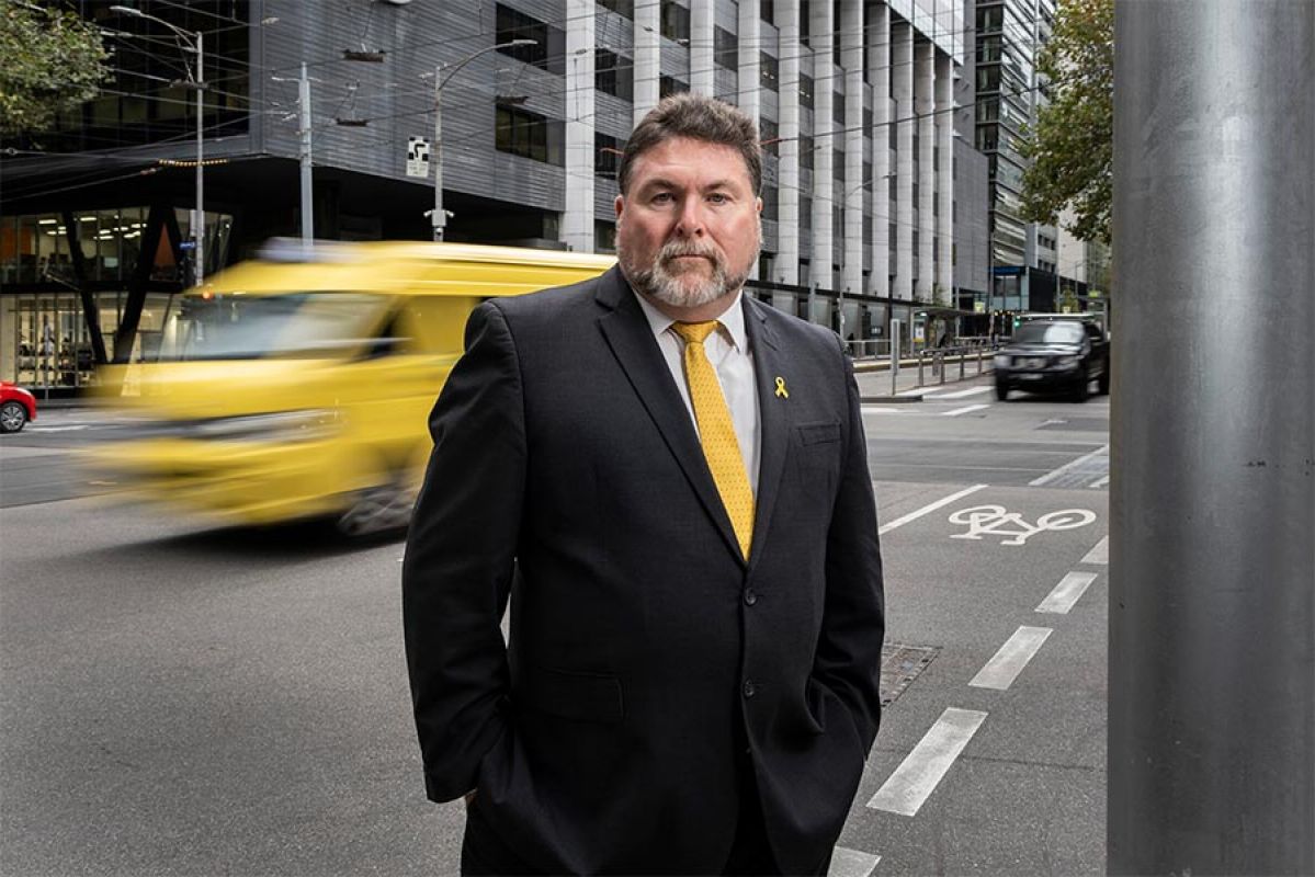 National Road Safety Week founder, Peter Frazer, standing on a street corner wearing a suit, yellow tie and matching yellow ribbon pin