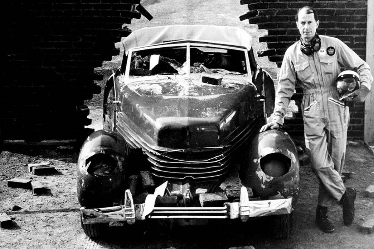 A black and white old photograph of a man with a crash test helmet leaning on an old-fashioned car that's just run through a brick wall