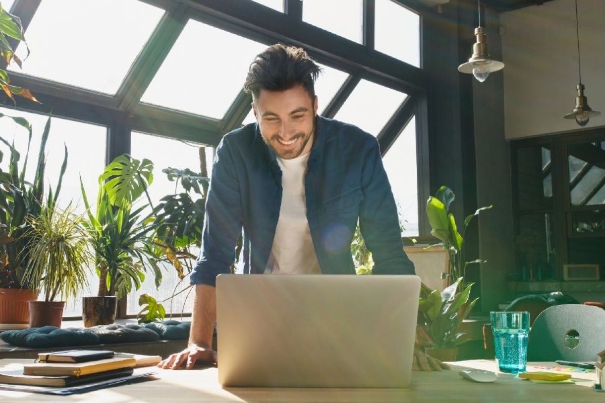 young man smiling at laptop in an office filled with sunlight and green potted plants