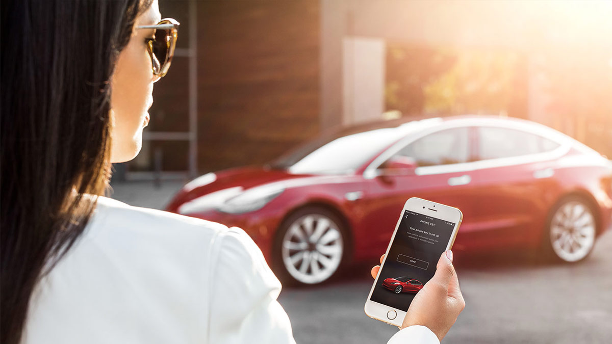 Woman holding a mobile phone with the Tesla app on screen, with a red Tesla Model 3 out of focus in the background