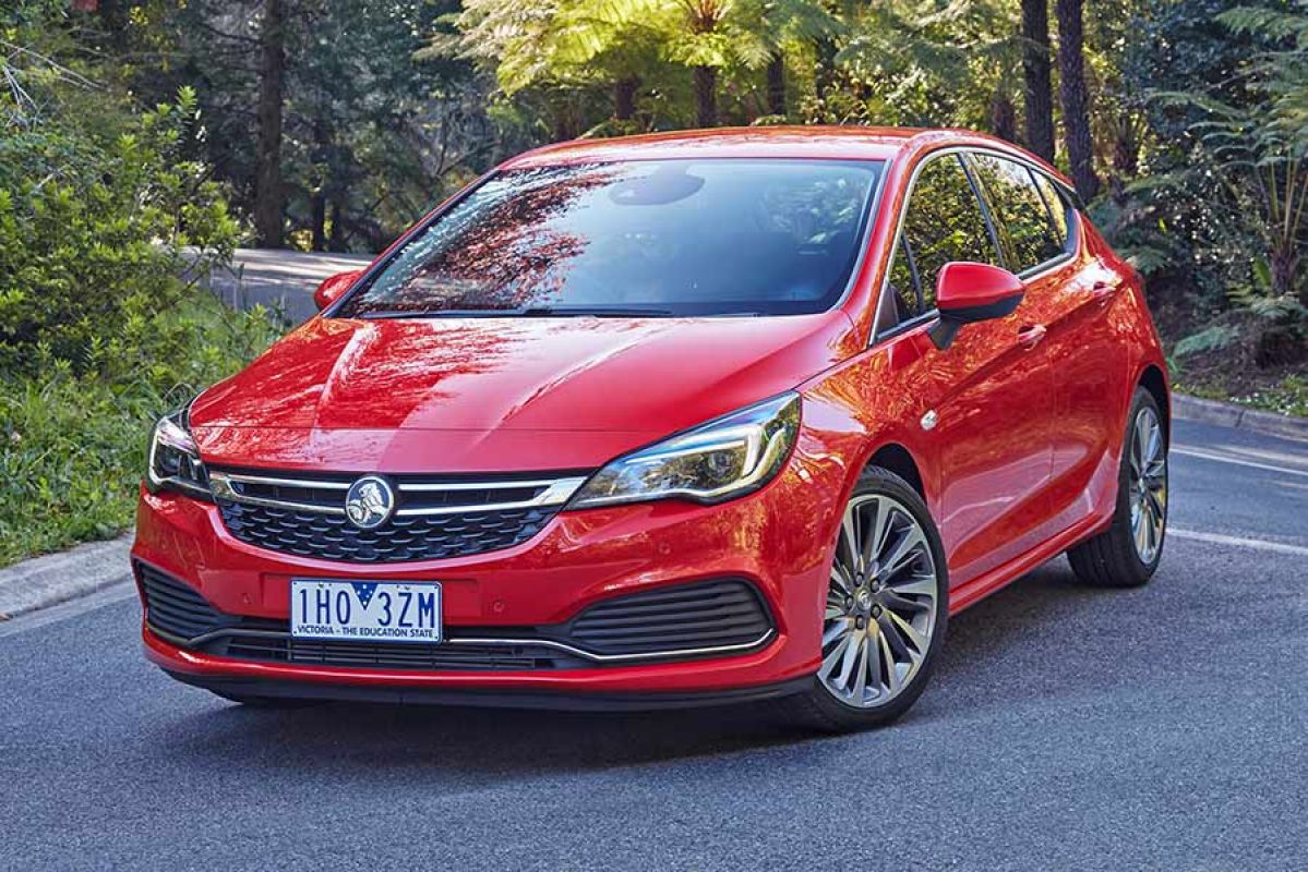 Front side view of a red Holden Astra 2017 car parked on a road flanked by a lush green forest