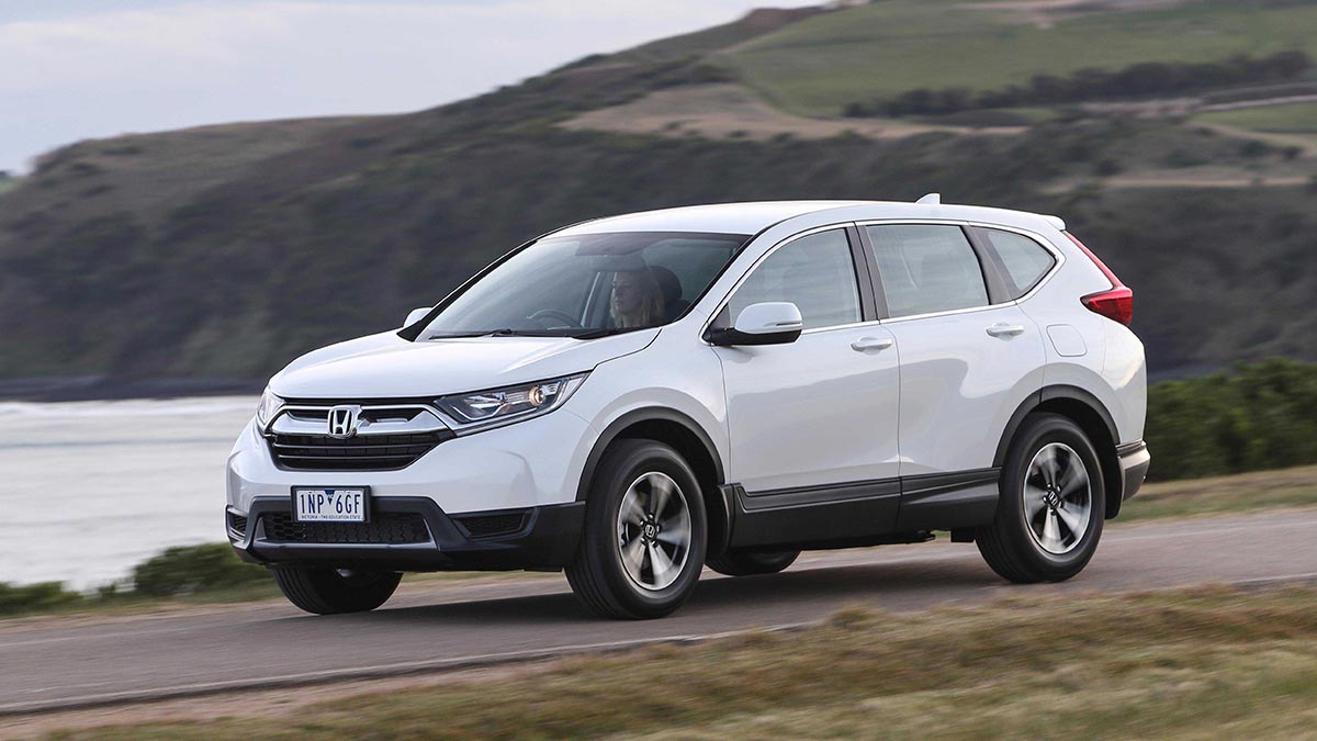 Front side view of a white Honda CR-V in motion on a dirt road next to a body of water