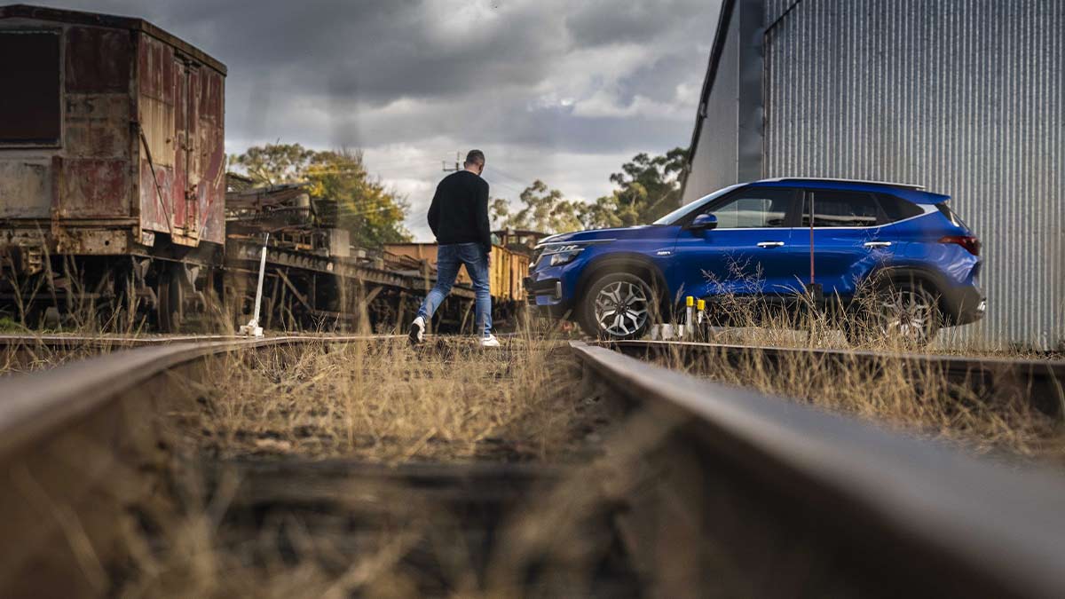 A man walking towards a blue Kia Seltos parked next to a train track