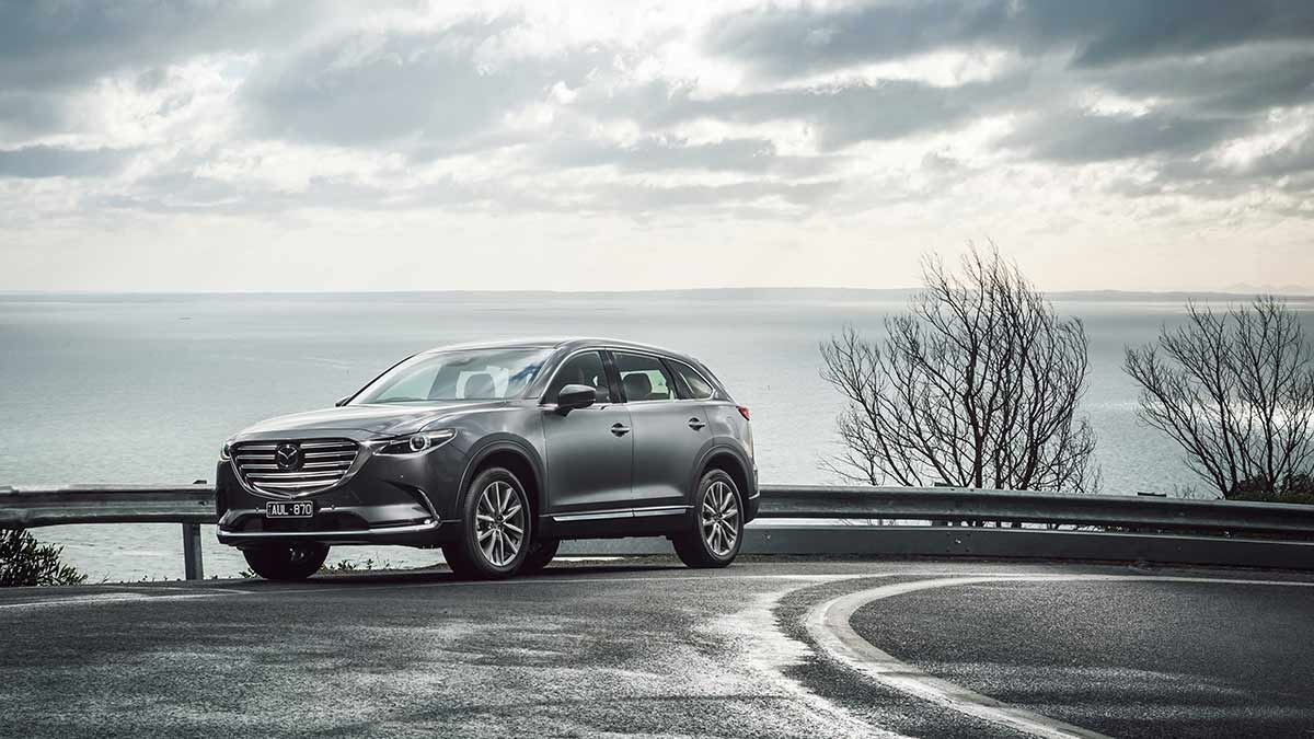 Front side view of a metallic grey Mazda CX-9 parked on the bend of a road next to the sea