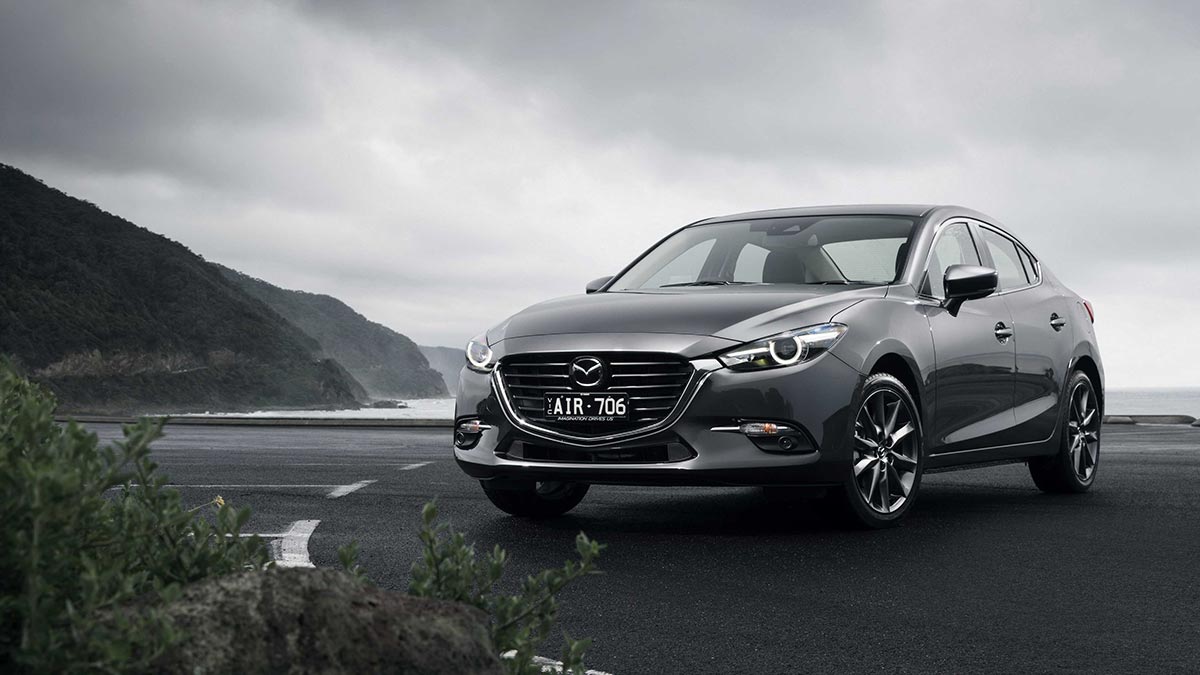 Front side view of a dark grey Mazda3 parked on tarmac next to the sea