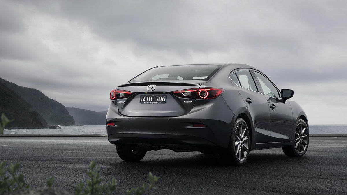 Rear side view of a dark grey Mazda3 parked on tarmac next to the sea