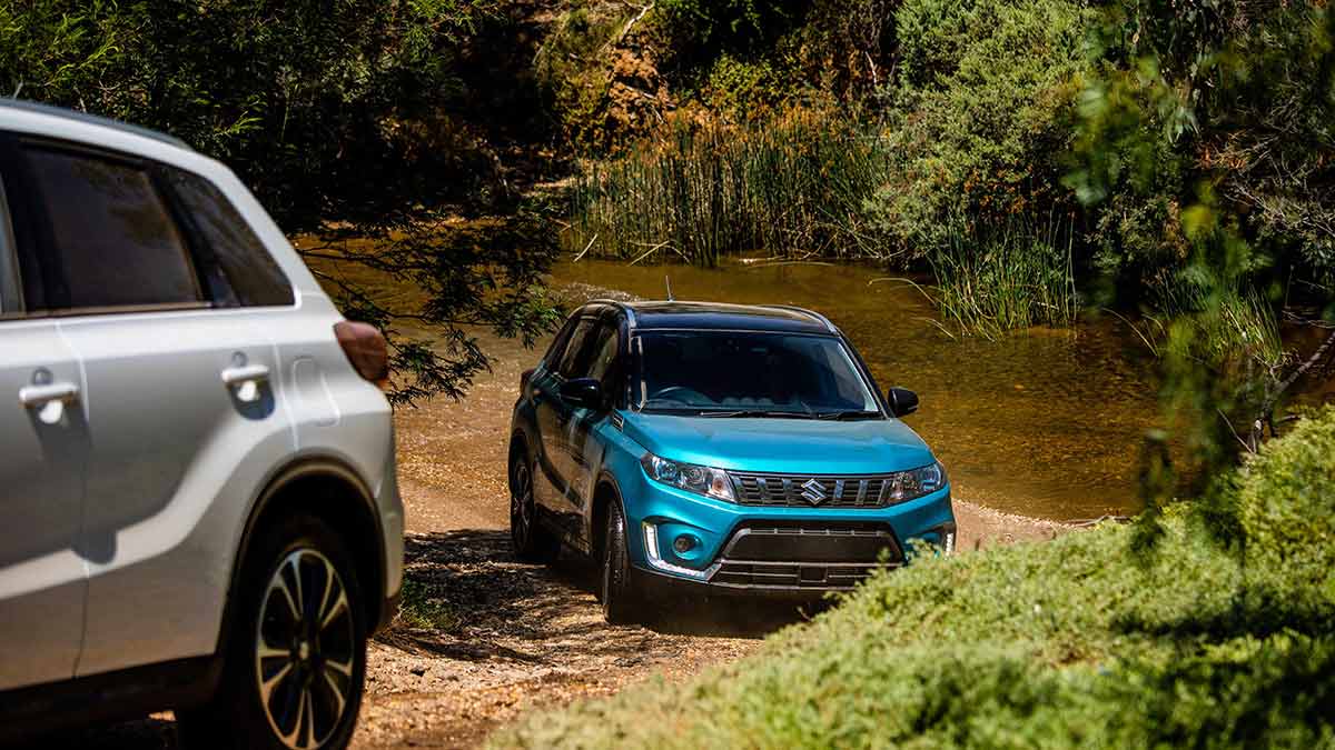 Front side view of a blue Suzuki Vitara AllGrip Turbo following a white Suzuki Vitara AllGrip Turbo through dirt and water