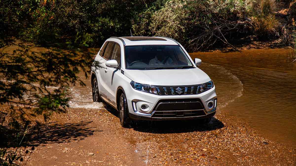 Front side view of a white Suzuki Vitara AllGrip Turbo driving through dirt and water