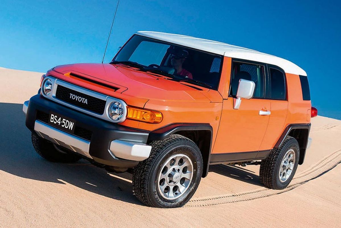 Front side view of an orange Toyota FJ Cruiser scaling a sand dune