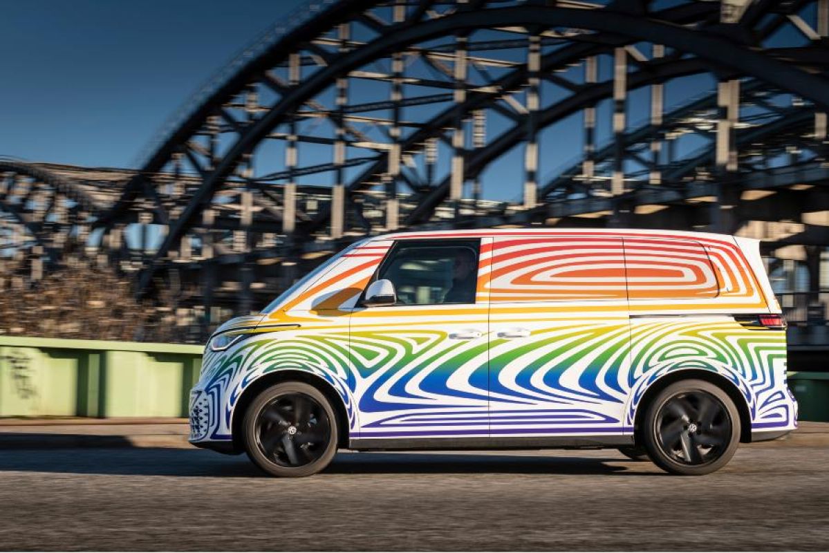 A rainbow-patterned Volkswagen ID.Buzz driving past a curved steel bridge