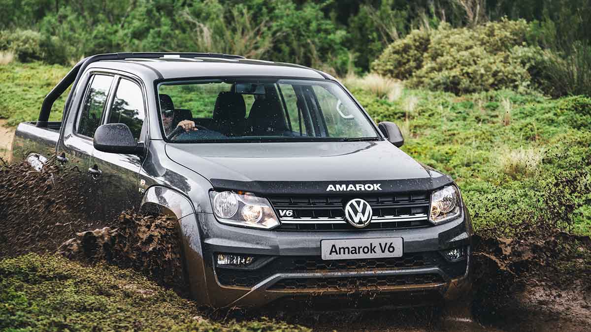 Front side view of a grey Volkswagen Amarok V6 driving through a muddy road in a green valley