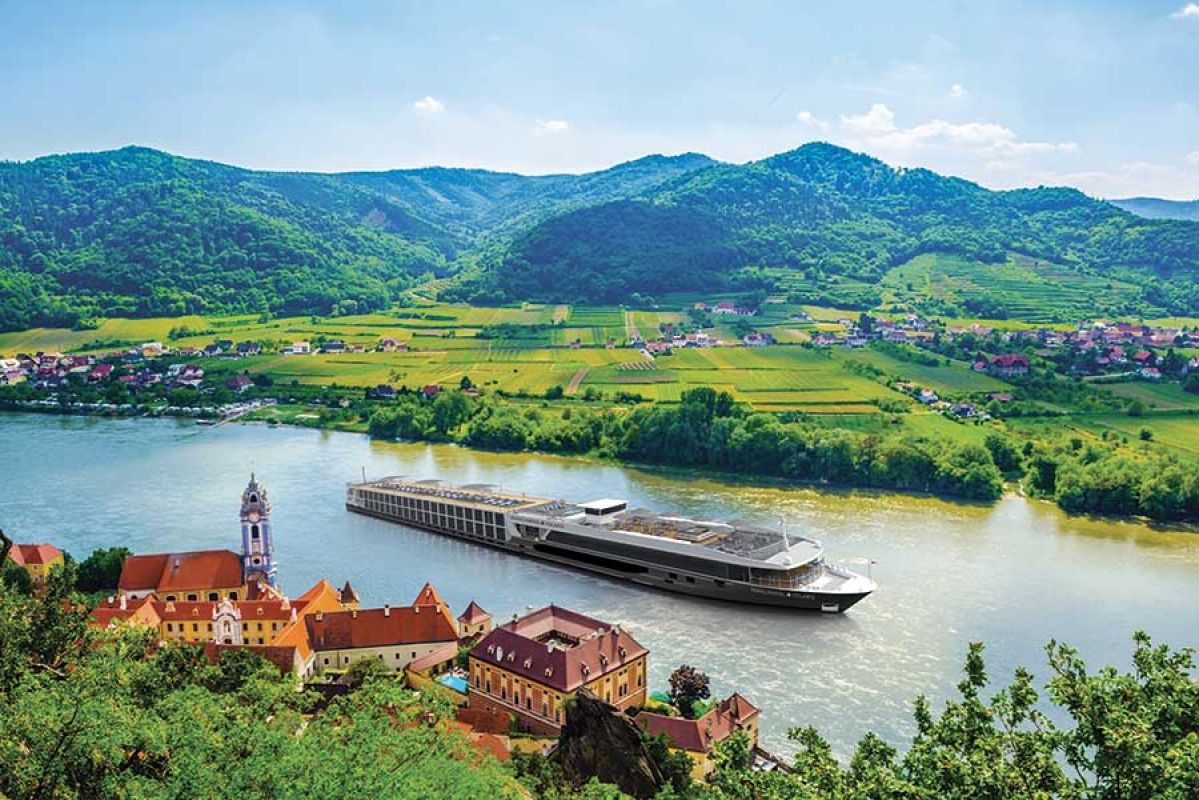 cruise ship going down a river with greenery and buildings in the background 