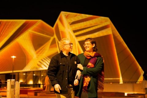 Two people walking past the Adelaide Festival Centre lit up for Illuminate Adelaide