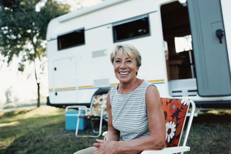A mature woman sits out front of her caravan smiling RACV Seniors Travel Insurance