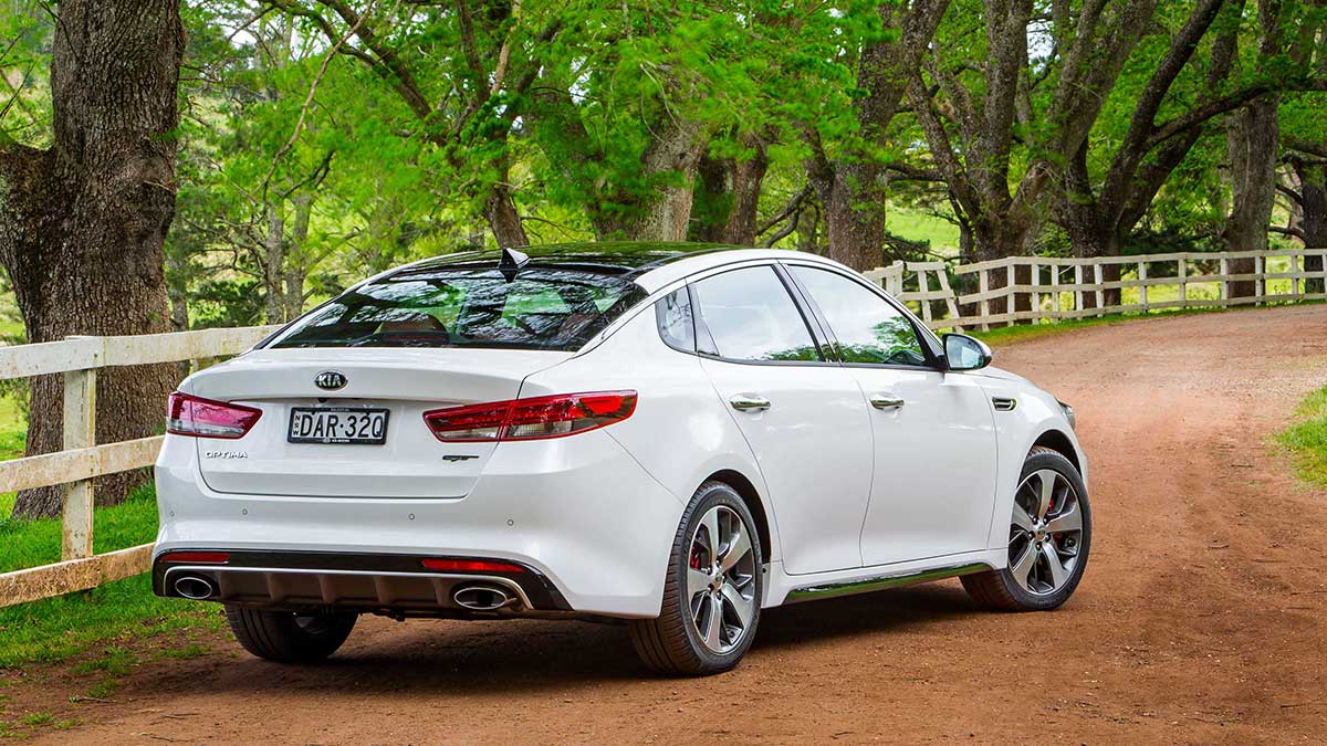 Rear side view of a white Kia Optima car parked on an unsealed road lined by trees