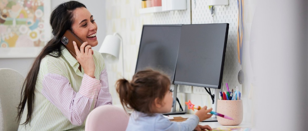 Woman on phone smiling in front of a monitor with her daughter drawing next to her Woman on phone smiling in front of a monitor with her daughter drawing next to her