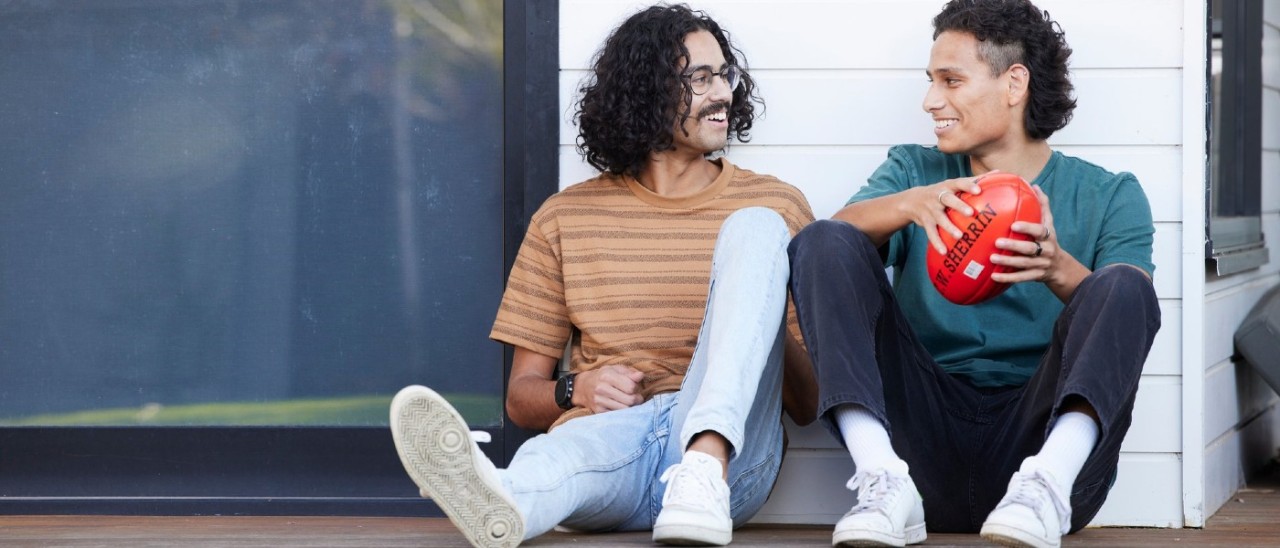 Young people Two young people sitting outside next to a white weatherboard house. They smile at each other and one holds an Aussie Rules football