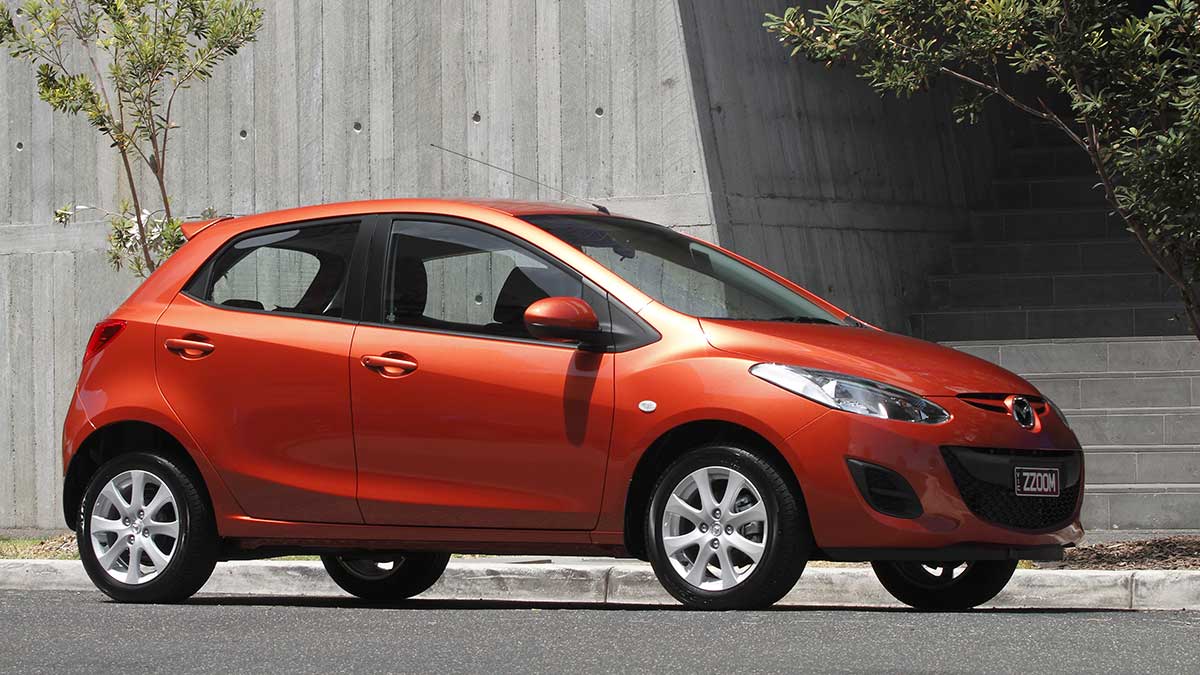 Front side view of a metallic orange Mazda2 parked in front of a building
