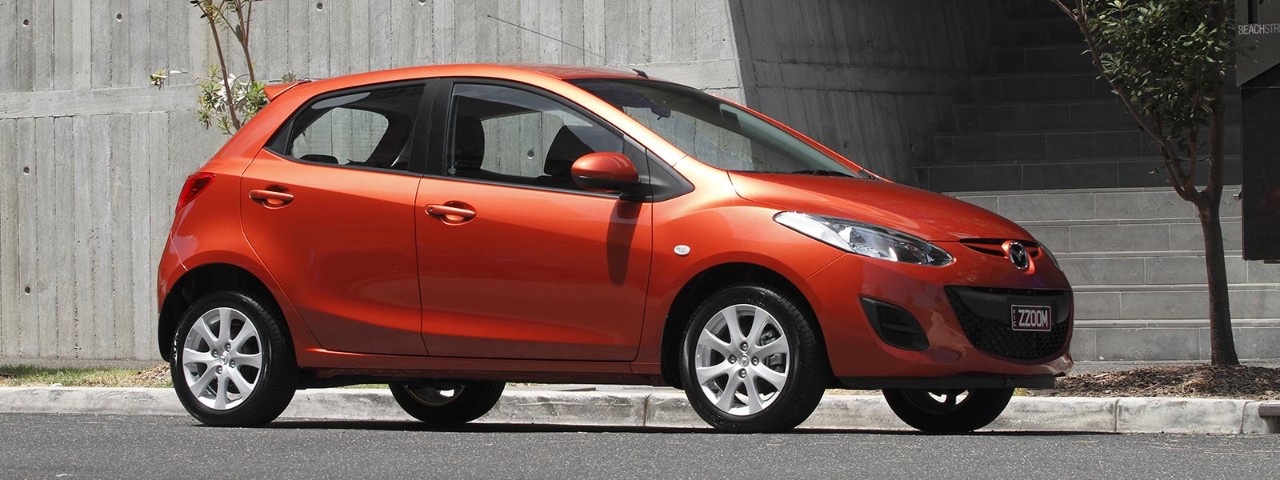 Front side view of a metallic orange Mazda2 parked in front of a building