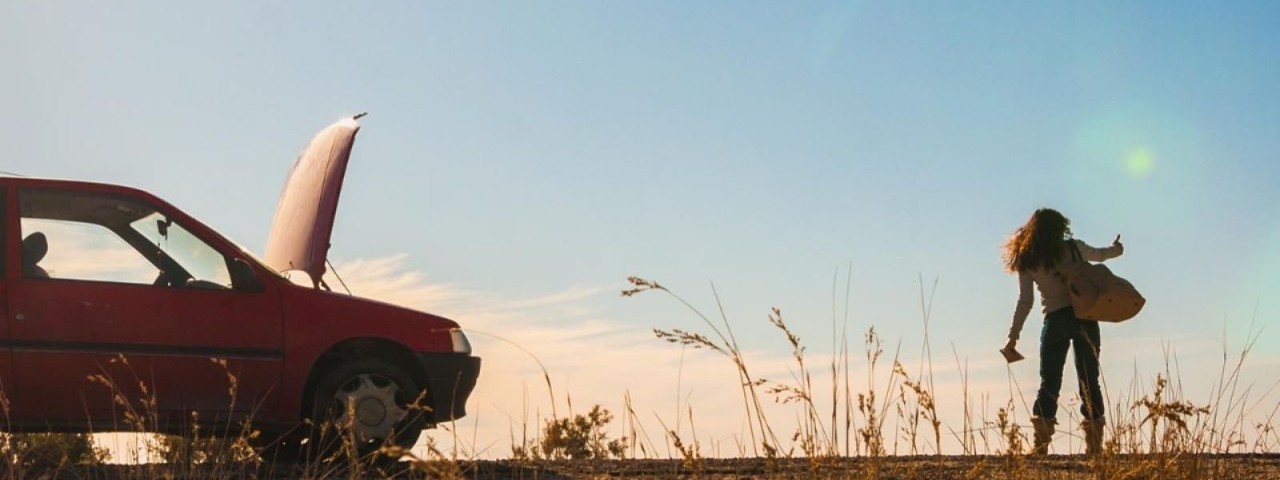 woman standing on highway trying to hitch hike next to car with bonnet up in field
