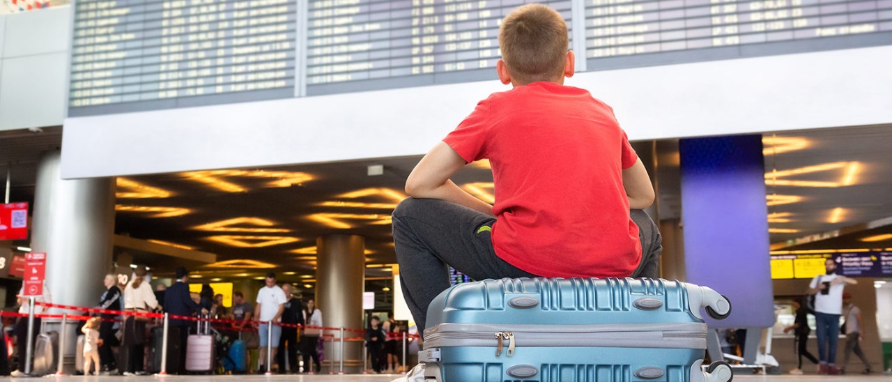 Flight-delay-GettyImages-1662197206-1400x600 Boy sitting on suitcase at airport looking at departures board